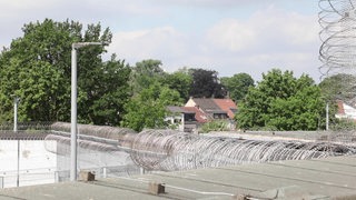 Man schaut über einen Drahtzaun hinweg auf eine Häuserreihe und Bäume.