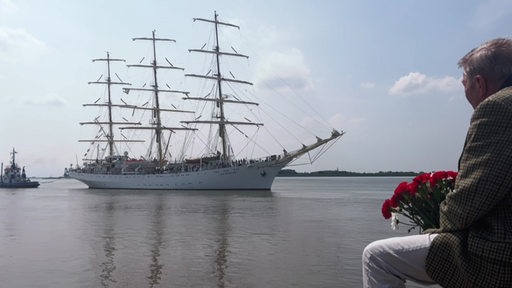 Ein Dreimaster läuft in den Hafen ein. Ein Mann sitzt mit Blumenstrauß wartend im Vordergrund.