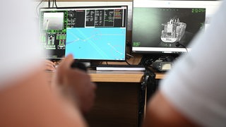 Zwei Personen sitzen mit einem Steuerknüppel vor zwei Bildschirmen worauf ein Schiff zu sehen ist.