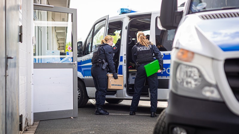 Eine Polizeibeamtin steht während einer Durchsuchung mit einem Karton an einem Polizeiwagen.