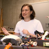 Eine Frau in einem weißen T-Shirt sitzt an einem Tisch, auf dem technische Bauteile liegen.