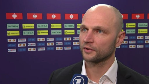 Eishockey-Manager Sebastian Furchner von den Fischtown Pinguins steht nach einem Champions-League-Spiel vor einer Werbewand beim Interview.