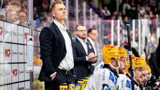 Alexander Sulzer, Cheftrainer des Eishockey-Teams der Fischtown Pinguins, steht mit ernster Miene an der Bande, vor ihm sitzen einige Ersatzspieler.