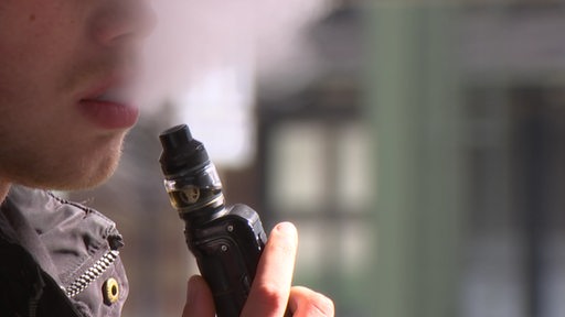 Ein Mann hält ein Vape in der Hand und pustet aus dem Mund Dampf aus.