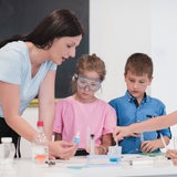 Eine Frau steht mit einem Jungen und zwei Mädchen an einem Tisch und zeigt ihnen ein kleines Fläschchen.