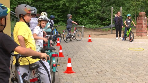 Mehrere Kinder stehen mit ihren Fahrrädern und Helmen aufgereiht hinter Verkehrshütchen.