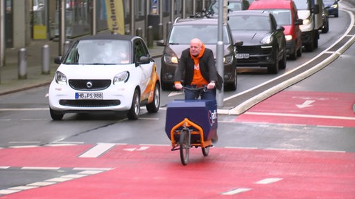 Ein Mann fährt mit seinem Lastenfahrrad auf einem breiten Radweg. Hinter ihm stehen mehrere Autos an einer Ampel.