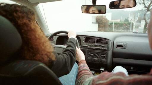 Der Fahrlehrer greift der Fahrschülerin korrigierend ins Lenkrad.