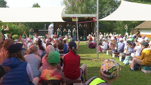 Viele Personen sitzen bei einem Ferienlager auf Bänken vor einer Bühne.