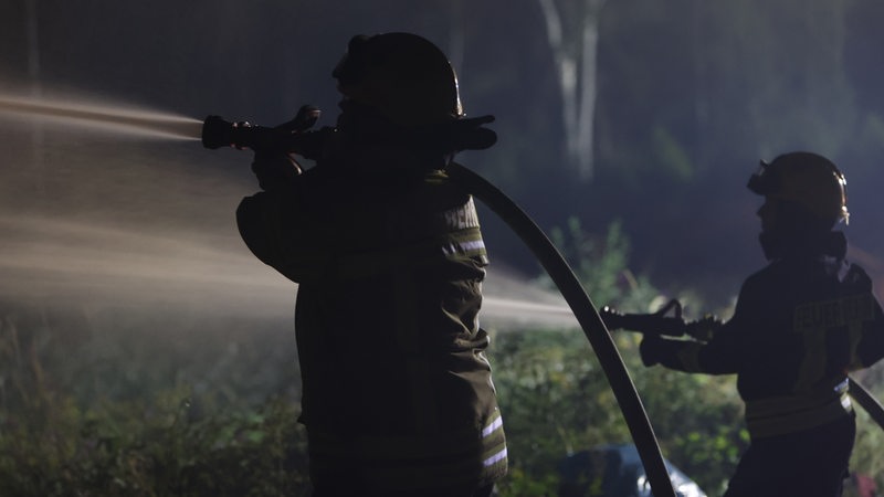 Zwei Einsatzkräfte der Feuerwehr löschen mit zwei Wasserschläuchen in der Nacht Feuer in einem Wald.