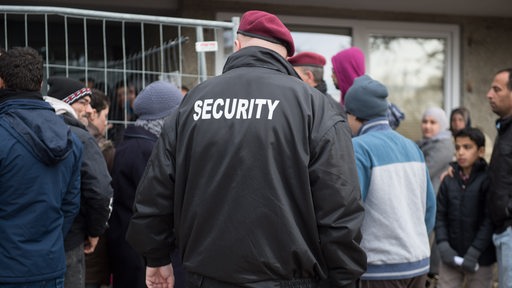 Rückenansicht eines Security-Mitarbeiters inmitten von Flüchtlingen vor einem Flüchtlingsheim