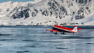 Ein Flugzeug fliegt übers Meer. Im Hintergrund ist ein Eisberg zu sehen.