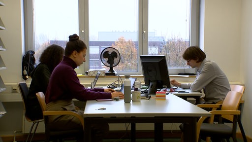 Mehrere Menschen sitzen an einem Schreibtisch vor Laptops.