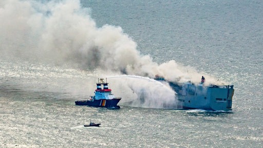 Ein Schiff richtet eine Wasserkanone auf einen brennenden Frachter. 