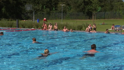 Ein Freibadbecken, in dem Leute schwimmen und am Beckenrand sitzen.