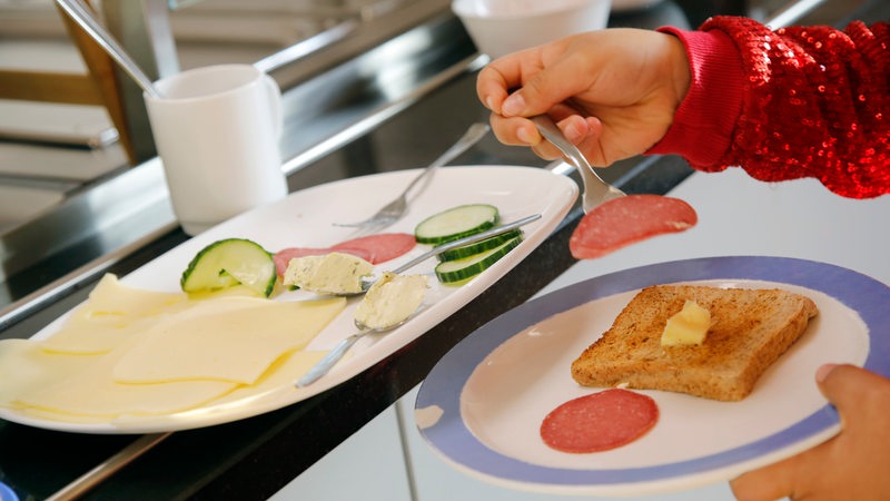 Eine Kinderhand lädt eine Scheibe Wurst auf einen Teller. Im Hintergrund sind Gurken- und Käsescheiben zu sehen.