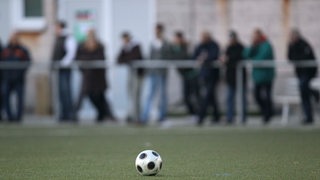 Zuschauer am Spielfeldrand unscharf im Hintergrund, im Vordergrund liegt ein Fußball auf dem Platz (Symbolbild)