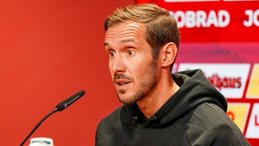 Freiburgs Fußball-Trainer Julian Schuster erklärt etwas mit weit geöffneten Augen bei einer Pressekonferenz.