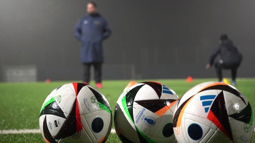 Wir sehen im Vordergrund 3 Fußballbälle auf einem Trainingsplatz.