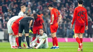 Werder-Stürmer Justin Njinmah hockt frustriert im Spiel gegen Bayern München auf dem Rasen, nachdem er umgeknickt ist. Der Schiedsrichter erkundigt sich bei ihm, auch andere Spieler beugen sich zu ihm herunter.