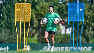 Werder-Profi Leonardo Bittencourt steht nachdenklich mit einem Ball unter jedem Arm nachdenklich am Rande des Trainings zwischen zwei großen Metallmännchen.