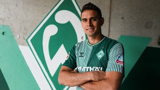 Werder-Neuzugang Rafael Borre posiert für ein Foto vor einer großen, aufgemalten Werder-Raute an der Wand.