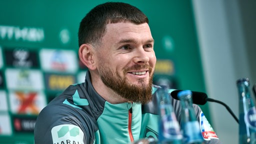 Werder-Spieler Oliver Burke sitzt bei einer Pressekonferenz am Mikrofon.