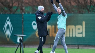 Werder-Stürmer Marvin Ducksch reißt am Rande des Trainings strahlend die Arme hoch, Trainer Ole Werner freut sich neben ihm und applaudiert.