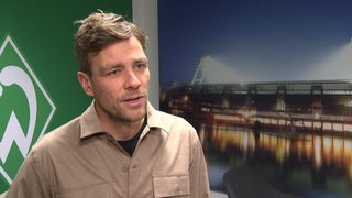 Werders Fußball-Geschäftsführer Clemens Fritz steht bei einem Interview vor einer Fototapete, auf dem das Weser-Stadion abgebildet ist.