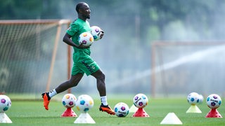 Werders Toptransfer Naby Keita rennt mit zwei Fußbällen in der Hand zwischen anderen Fußbällen auf dem Rasen entlang.