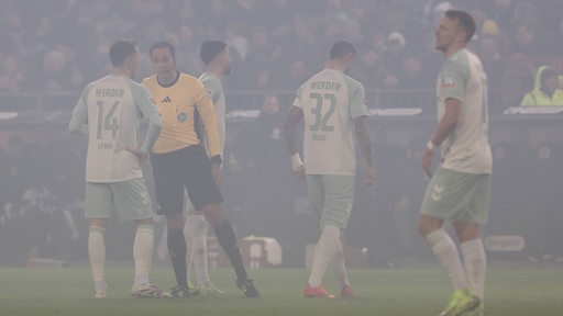 Werder-Spieler stehen im dicken Nebel durch Pyrotechnik auf dem Spielfeld und warten auf die Entscheidung des Schiedsrichters.