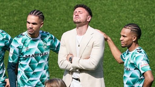 Werder-Profi Milos Veljkovic steht im hellen Anzug neben seinen Teamkollegen auf dem Spielfeld bei seiner Verabschiedung, die Arme vor sich verschränkt, den Kopf in den Nacken gelegt und sichtlich ergriffen.