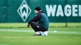 Werder-Trainer Ole Werner trägt eine schwarze Wollmütze und hockt am Rande des Trainings auf einem Fußball, schaut ins Leere.