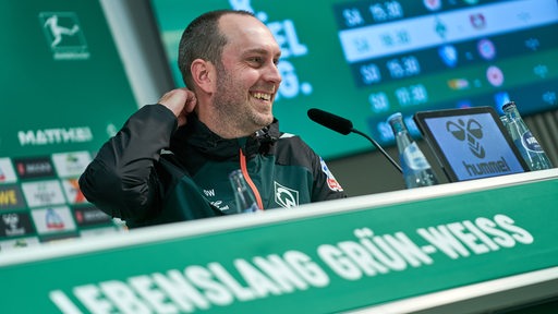 Werder-Trainer Ole Werner sitzt lachend bei einer Pressekonferenz.