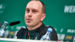 Werder-Trainer Ole Werner sitzt lächelnd auf dem Podium bei einer Pressekonferenz.