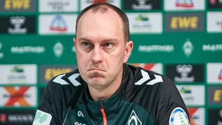 Werder-Trainer Ole Werner sitzt auf dem Podium einer Pressekonferenz und verzieht nachdenklich das Gesicht.