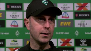 Werder-Trainer Ole Werner steht nach dem Spiel gegen Stuttgart vor einer Werbewand beim Interview.