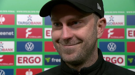 Werder-Trainer Ole Werner steht nach dem Pokalspiel gegen Darmstadt lächelnd vor einer Werbewand beim Interview.