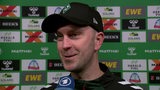 Werder-Trainer Ole Werner steht lächelnd nach dem Spiel vor einer Werbewand beim Interview.