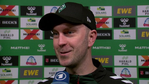 Werder-Trainer Ole Werner steht lächelnd nach dem Spiel vor einer Werbewand beim Interview.