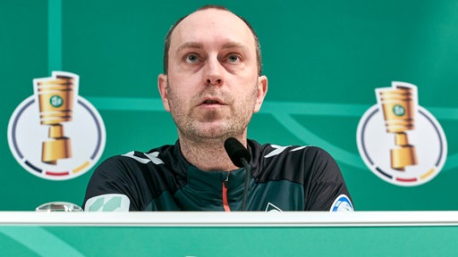 Ole Werner sitzt bei einer Pressekonferenz. Links und rechts hinter ihm ist der DFB-Pokal abgebildet.