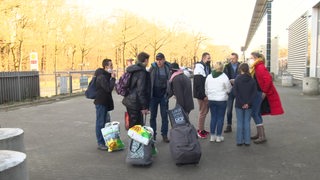 Eine Gruppe von Menschen steht in einem losen Kreis. Sie haben gepackte Taschen und Plastiktüten dabei.