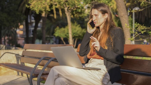 Eine junge Geschäftsfrau sitzt mit ihrem Laptop telefonierend auf einer Parkbank.