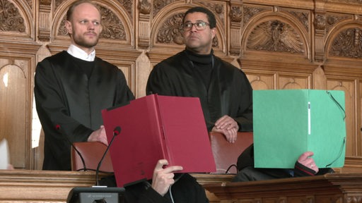 Zwei angeklagte Personen verdecken sich mit Mappen das Gesicht in einem Gerichtssaal