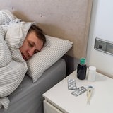 Ein Mann liegt krank im Bett. auf dem Nachttisch liegen ein Fieberthermometer und Tabletten.