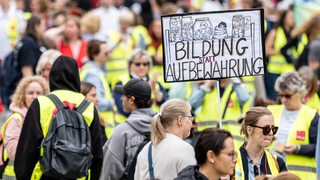 Menschen nehmen an einer Kundgebung der Gewerkschaften GEW und Verdi teil. Auf einem Schild ist die Aufschrift "Bildung statt Aufbewahrung" zu lesen.