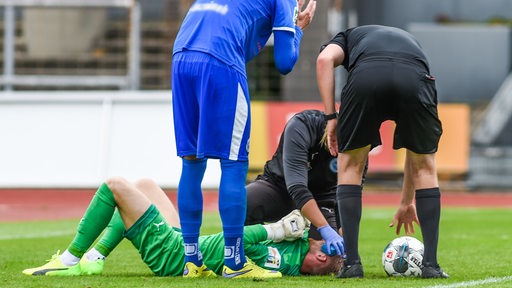 Ein Torwart liegt nach einem Foulspiel verletzt am Boden. Schiedsrichter, Sanitäter und ein Spieler stehen daneben.