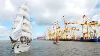 Ein großes und ein kleines Segelschiff fahren an Kränen eines Hafens entlang.