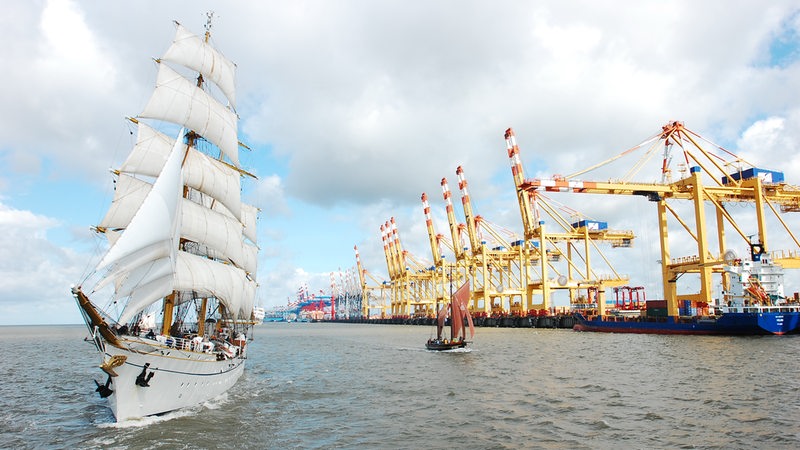 Ein großes und ein kleines Segelschiff fahren an Kränen eines Hafens entlang.
