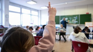 Eine Schülerin hebt die Hand. während der Lehrer an der Tafel schreibt.